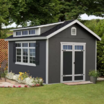 Do-it Yourself Windemere A gray garden shed with white trim, double doors, and windows is situated in a green yard near a small patio area with flowers, foliage, and outdoor furniture.