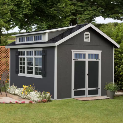 Do-it Yourself Windemere A gray garden shed with white trim, double doors, and windows is situated in a green yard near a small patio area with flowers, foliage, and outdoor furniture.