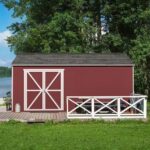 A red shed stands beside a white fence, featuring a wooden deck in a serene outdoor setting